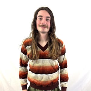 May include: A vintage-style, long-sleeved sweater with a V-neck. The sweater features horizontal stripes in shades of brown, beige, and orange. The bottom hem and cuffs are a darker brown. The person is wearing olive green pants.