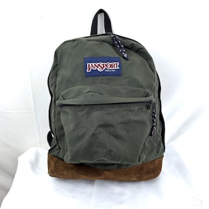 May include: A green Jansport backpack with a brown suede bottom and a front pocket. The backpack has a black handle and zipper pulls with a blue and white cord. The Jansport logo is on the front.