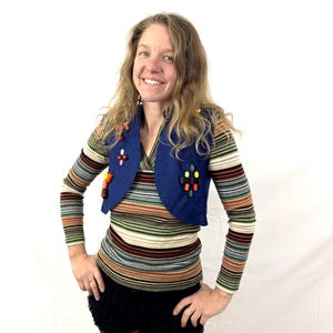 May include: A woman wearing a blue vest with colorful embellishments over a striped long-sleeve shirt and black shorts. The vest has a rounded shape and is decorated with various colored beads and shapes. The shirt features horizontal stripes in multiple colors.