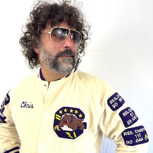 May include: Cream-colored varsity jacket with purple and white striped accents. Features a large patch with a bulldog graphic and the name "Chris" embroidered on the chest. Additional patches on the sleeves display text including "REG. CHAM" and numbers.