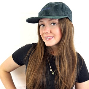 May include: A woman with long brown hair is wearing a black t-shirt and a green and black plaid baseball cap with the word "Wilson" embroidered on the front. She is smiling and looking at the camera.