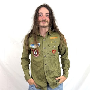 May include: An olive green long-sleeve button-down shirt featuring multiple embroidered patches on the chest and sleeves, including a circular red and black emblem, a rectangular orange patch, and a small red heart-shaped patch on the pocket.