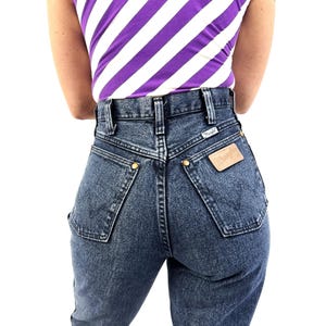 Puede incluir: Vaqueros de cintura alta, de color azul oscuro, con una etiqueta Wrangler. Los vaqueros tienen un dise&ntilde;o cl&aacute;sico de cinco bolsillos con remaches dorados y un parche de cuero. La persona lleva una camiseta de rayas moradas y blancas.