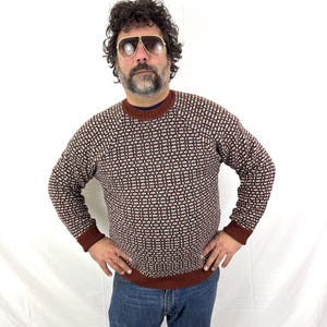 Puede incluir: Un su&eacute;ter con estampado marr&oacute;n y blanco con cuello redondo y mangas largas. El su&eacute;ter tiene pu&ntilde;os marrones y un dobladillo marr&oacute;n. La persona de la imagen lleva vaqueros azules y gafas de sol de aviador.