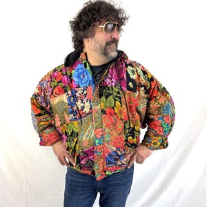 Op de afbeelding: Een kleurrijke gewatteerde jas met een patchwork ontwerp met levendige bloempatronen in rood, blauw, geel en groen. De jas heeft een zwarte capuchon en een ritssluiting. De mouwen zijn gerimpeld en het wordt gedragen over een donker topje en een blauwe jeans.