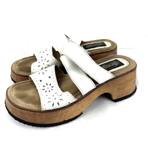 May include: White leather sandals with a floral cut-out design and wooden platform heels. The sandals have a brown sole and the brand name "Bongo" is visible on the insole. These vintage sandals are a stylish footwear option.