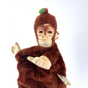 May include: A brown plush monkey puppet with a green and red pom-pom on its head. The monkey has a brown fur coat and a light brown face with a painted expression. The puppet is holding its arms in front of it.