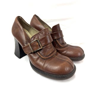 May include: A pair of brown leather heeled shoes with a buckle closure. The shoes have a chunky heel and a rounded toe.