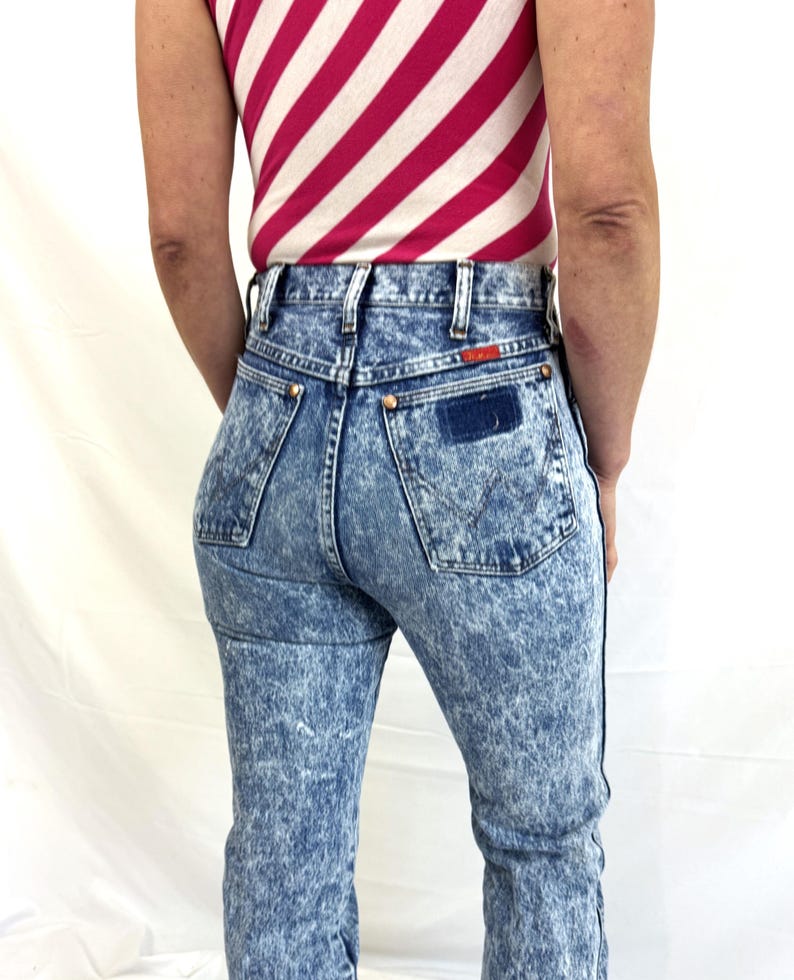 Puede incluir: Vaqueros de mezclilla con lavado &aacute;cido, de cintura alta y pernera recta. Los vaqueros tienen un efecto marmoleado azul y blanco, con un parche azul oscuro en el bolsillo trasero. La persona lleva una camiseta sin mangas a rayas rosas y blancas.