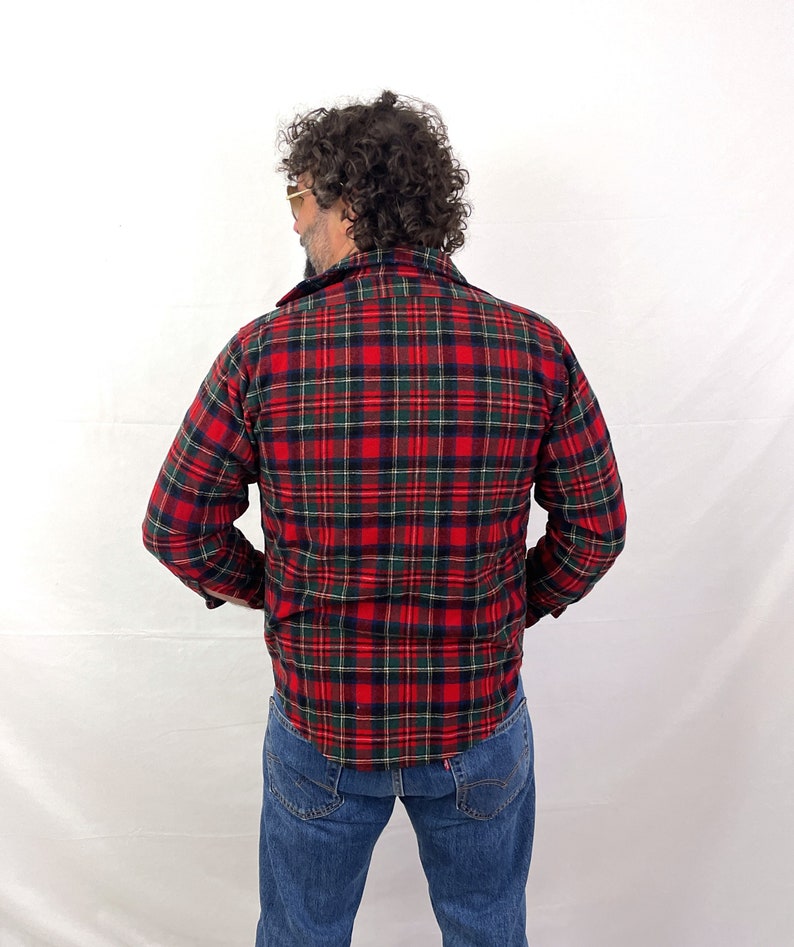 May include: A red, green, and black plaid flannel shirt with long sleeves. The shirt is buttoned up and the collar is turned up.