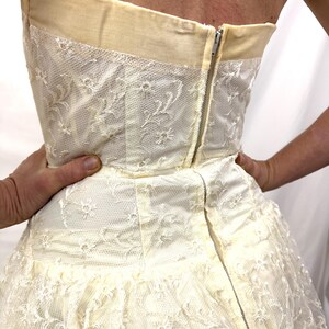 Puede incluir: Vestido marfil sin tirantes con una delicada superposici&oacute;n de encaje floral. El vestido presenta un corpi&ntilde;o ajustado con un ribete color crema y una falda amplia. Una cremallera plateada recorre la espalda. El vestido es sostenido por manos.