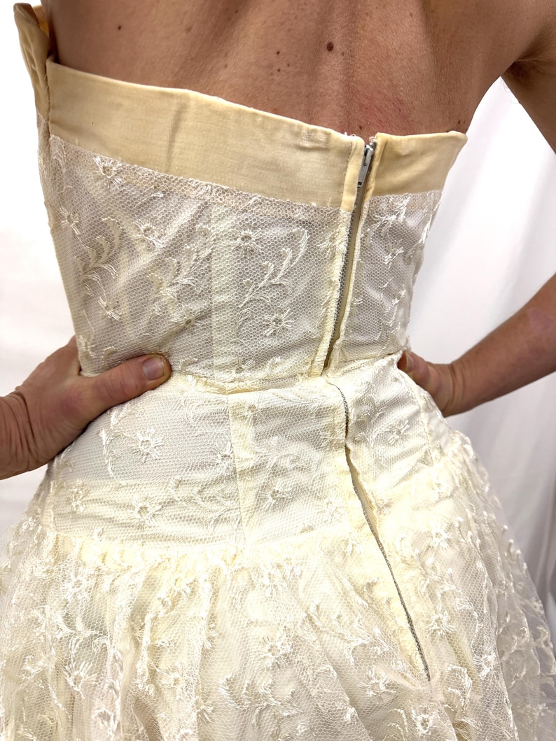 Puede incluir: Vestido marfil sin tirantes con una delicada superposici&oacute;n de encaje floral. El vestido presenta un corpi&ntilde;o ajustado con un ribete color crema y una falda amplia. Una cremallera plateada recorre la espalda. El vestido es sostenido por manos.