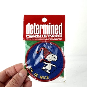 Puede incluir: Un parche redondo de Peanuts con Snoopy esquiando, con las palabras "ICE IS NICE". El parche es azul con detalles rojos y blancos, y está empaquetado en una funda de plástico transparente con un encabezado rojo que dice "determined PEANUTS PATCH".
