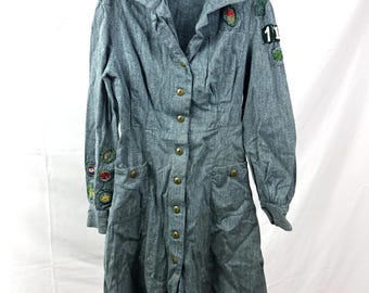 May include: A gray denim dress with long sleeves, button-up front, and pockets. The dress features embroidered patches on the sleeves and chest, and is hanging on a clothesline with wooden clothespins. The dress has a vintage aesthetic.