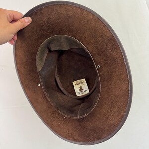 Puede incluir: Un sombrero de ante marr&oacute;n con ala ancha y una banda interior de color marr&oacute;n oscuro. El interior presenta una etiqueta blanca con el texto "SQUASHY" y "GRIZZLY MADE IN AUSTRALIA". Dos peque&ntilde;os ojales met&aacute;licos plateados son visibles en el ala.