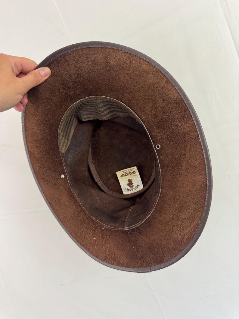 Puede incluir: Un sombrero de ante marr&oacute;n con ala ancha y una banda interior de color marr&oacute;n oscuro. El interior presenta una etiqueta blanca con el texto "SQUASHY" y "GRIZZLY MADE IN AUSTRALIA". Dos peque&ntilde;os ojales met&aacute;licos plateados son visibles en el ala.