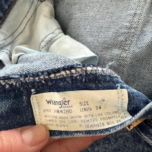 Puede incluir: Primer plano de unos vaqueros de mezclilla azules. La etiqueta de la marca Wrangler es visible, mostrando el estilo, la talla 9 y la longitud 34. Los vaqueros est&aacute;n hechos en EE. UU. La tela tiene una apariencia texturizada.