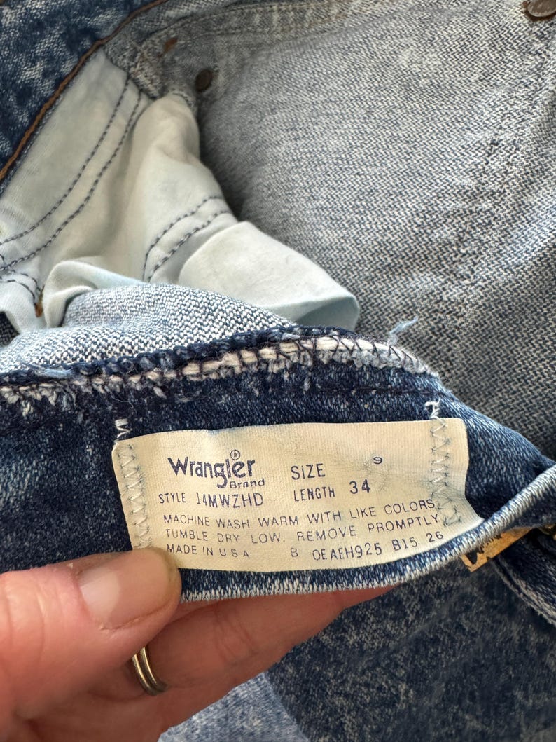 Puede incluir: Primer plano de unos vaqueros de mezclilla azules. La etiqueta de la marca Wrangler es visible, mostrando el estilo, la talla 9 y la longitud 34. Los vaqueros est&aacute;n hechos en EE. UU. La tela tiene una apariencia texturizada.