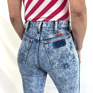 Puede incluir: Vaqueros de mezclilla con lavado &aacute;cido, de cintura alta y dise&ntilde;o cl&aacute;sico de cinco bolsillos. Los vaqueros tienen un parche Wrangler en el bolsillo trasero y remaches color lat&oacute;n. La modelo lleva un top a rayas rosas y blancas.