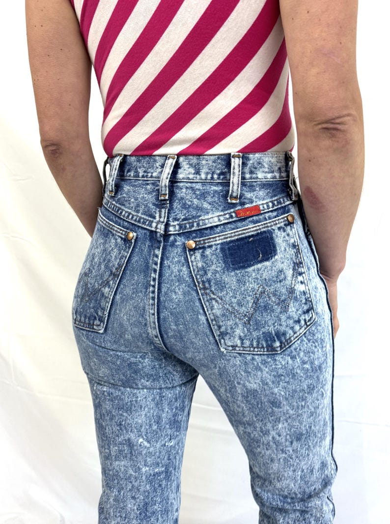 Puede incluir: Vaqueros de mezclilla con lavado &aacute;cido, de cintura alta y dise&ntilde;o cl&aacute;sico de cinco bolsillos. Los vaqueros tienen un parche Wrangler en el bolsillo trasero y remaches color lat&oacute;n. La modelo lleva un top a rayas rosas y blancas.
