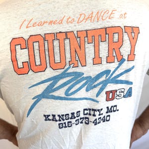 Puede incluir: Una camiseta gris claro con el texto "I Learned to DANCE at COUNTRY ROCK USA" en naranja y azul. La camiseta tambi&eacute;n tiene el texto "KANSAS CITY, MO. 816-373-4240".