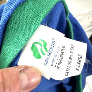 Puede incluir: Camiseta azul y verde de las Girl Scouts con una etiqueta que dice "GIRL SCOUTS" y "MADE IN U.S.A.". La etiqueta tambi&eacute;n incluye el n&uacute;mero de cat&aacute;logo 8-617 y la talla X-LARGE.