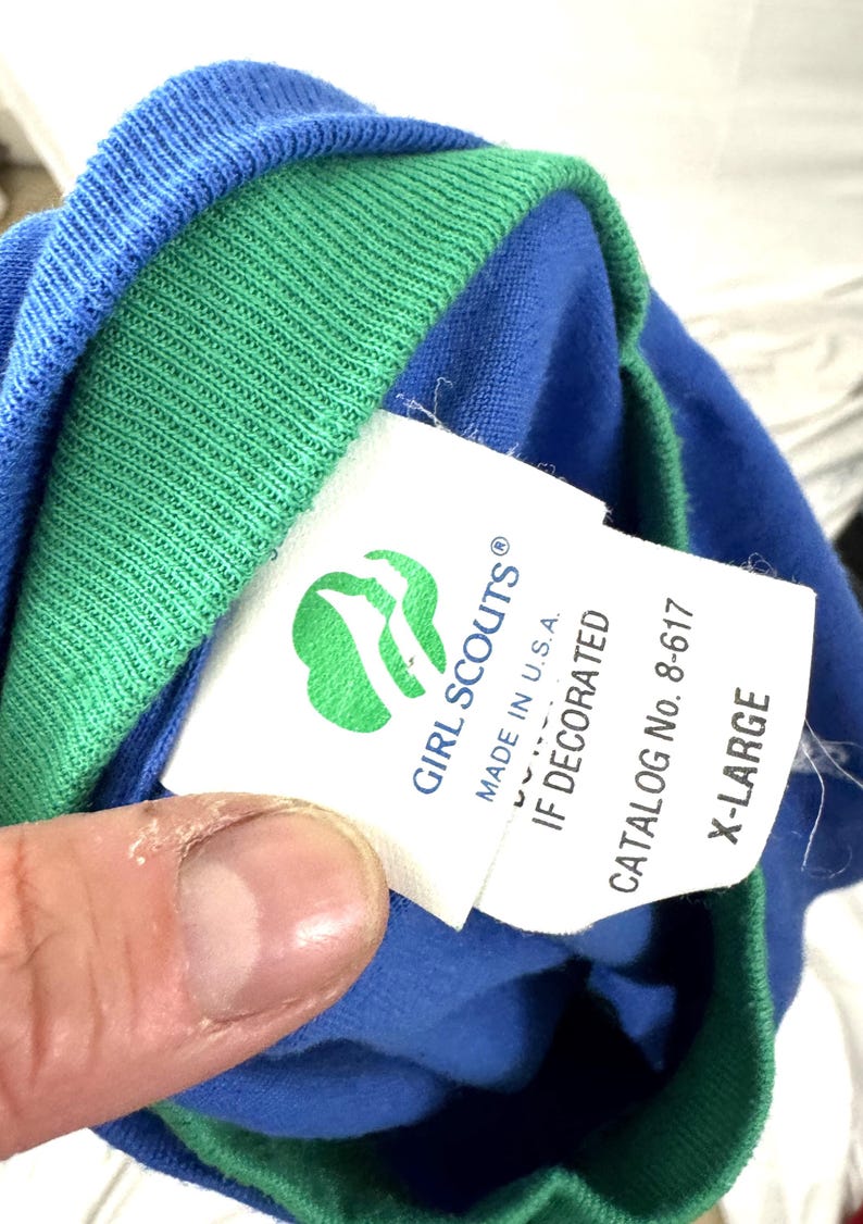 Puede incluir: Camiseta azul y verde de las Girl Scouts con una etiqueta que dice "GIRL SCOUTS" y "MADE IN U.S.A.". La etiqueta tambi&eacute;n incluye el n&uacute;mero de cat&aacute;logo 8-617 y la talla X-LARGE.