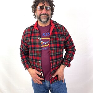 May include: A man wearing a red and black plaid flannel shirt over a maroon t-shirt with the text "King Fest" printed on it. He is also wearing blue jeans and aviator sunglasses.