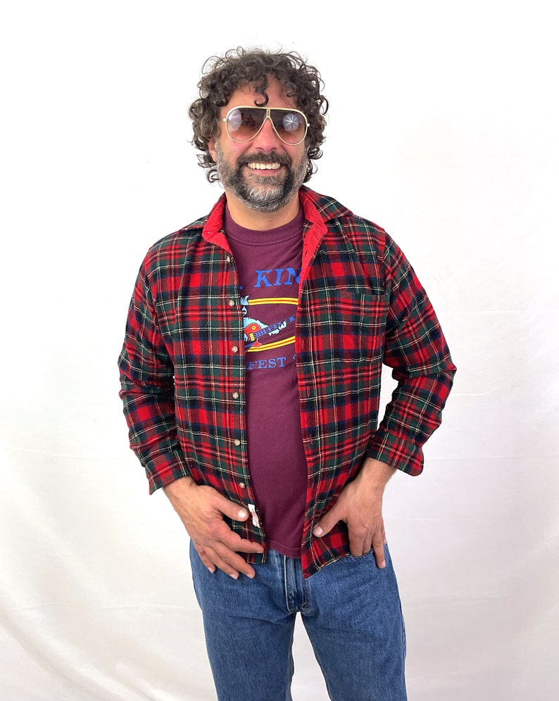 May include: A man wearing a red and black plaid flannel shirt over a maroon t-shirt with the text "King Fest" printed on it. He is also wearing blue jeans and aviator sunglasses.