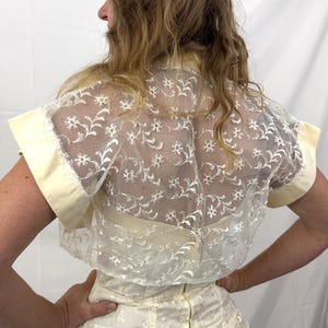 Puede incluir: Top de encaje marfil con motivos florales y mangas cortas. El top tiene una superposici&oacute;n transparente y se combina con una falda a juego. Las mangas y el cuello est&aacute;n ribeteados con una tela amarillo p&aacute;lido. La parte trasera del top es visible.