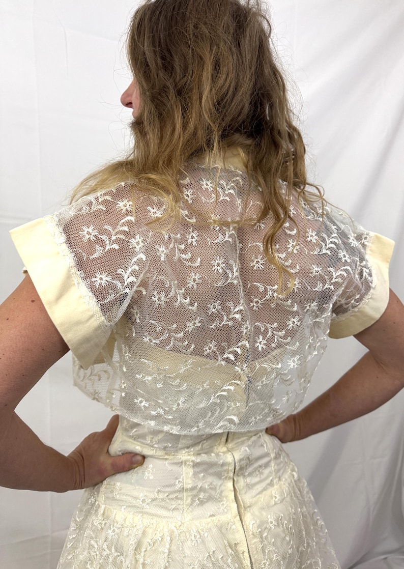 Puede incluir: Top de encaje marfil con motivos florales y mangas cortas. El top tiene una superposici&oacute;n transparente y se combina con una falda a juego. Las mangas y el cuello est&aacute;n ribeteados con una tela amarillo p&aacute;lido. La parte trasera del top es visible.