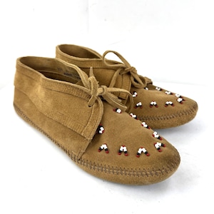 May include: A pair of tan suede moccasin shoes with decorative beadwork. The shoes feature a lace-up design and are adorned with small red, white, and black beads. The soles are stitched with a visible thread.