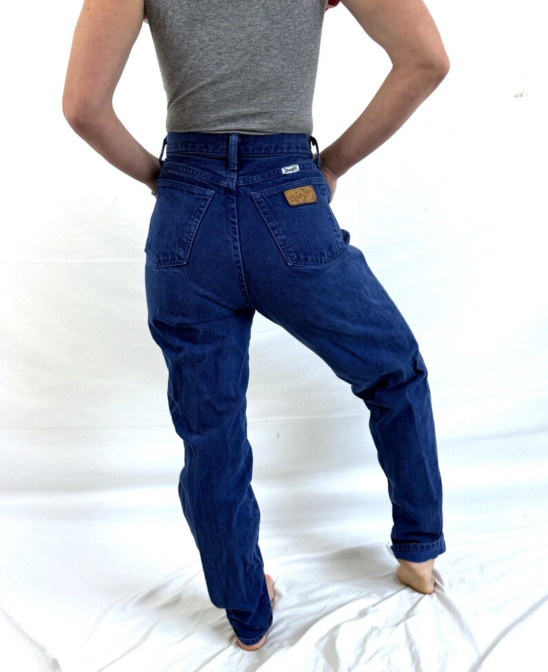 K&ouml;nnte beinhalten: Eine blaue Jeans mit hoher Taille und einem braunen Leder-Wrangler-Etikett auf der Ges&auml;&szlig;tasche.