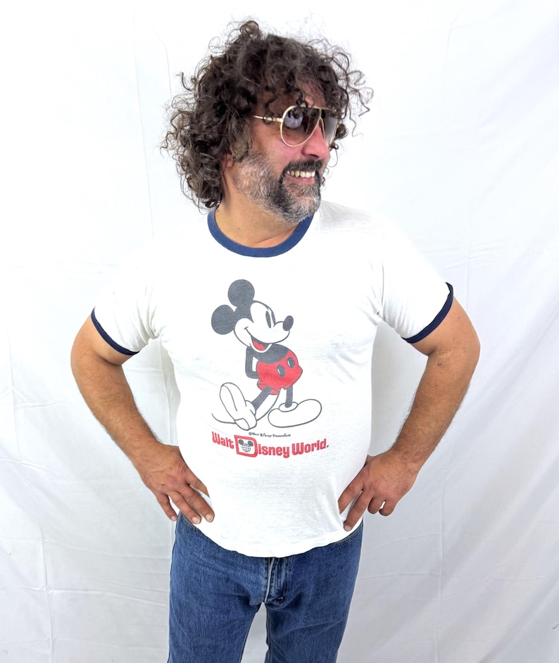 May include: White ringer t-shirt featuring a graphic of Mickey Mouse with red shorts and the text "Walt Disney World." The shirt has navy blue trim around the collar and sleeves. The person is wearing blue jeans and gold-rimmed sunglasses.