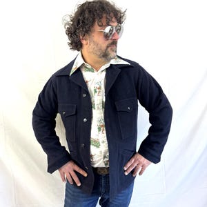 Puede incluir: Chaqueta azul marino con dos bolsillos en el pecho y dos bolsillos inferiores. La chaqueta est&aacute; abotonada y se lleva sobre una camisa estampada. La persona lleva vaqueros azules y gafas de sol.