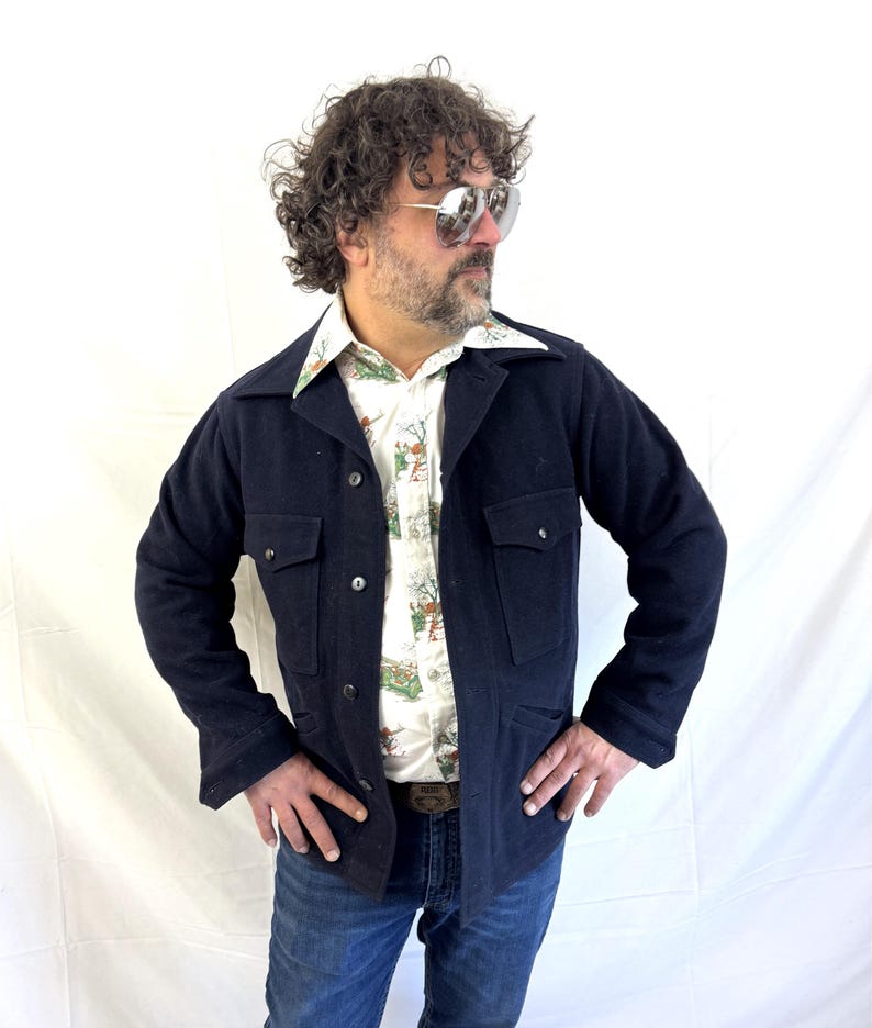 Puede incluir: Chaqueta azul marino con dos bolsillos en el pecho y dos bolsillos inferiores. La chaqueta est&aacute; abotonada y se lleva sobre una camisa estampada. La persona lleva vaqueros azules y gafas de sol.