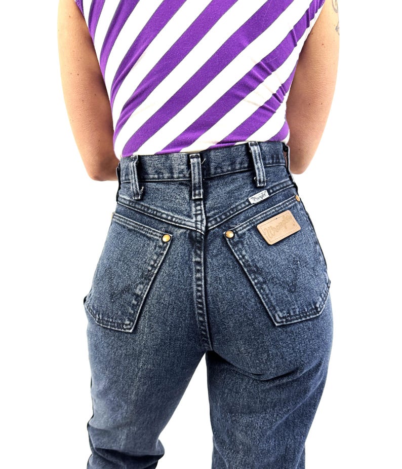 Puede incluir: Vaqueros de cintura alta, de color azul oscuro, con una etiqueta Wrangler. Los vaqueros tienen un dise&ntilde;o cl&aacute;sico de cinco bolsillos con remaches dorados y un parche de cuero. La persona lleva una camiseta de rayas moradas y blancas.