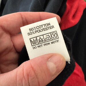 May include: A white clothing tag with black text and care symbols is held up. The tag reads "50% COTTON 50% POLYESTER" and includes washing instructions. The background shows a black and red garment.