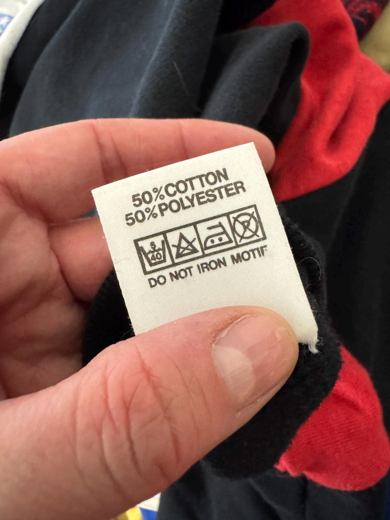 May include: A white clothing tag with black text and care symbols is held up. The tag reads "50% COTTON 50% POLYESTER" and includes washing instructions. The background shows a black and red garment.