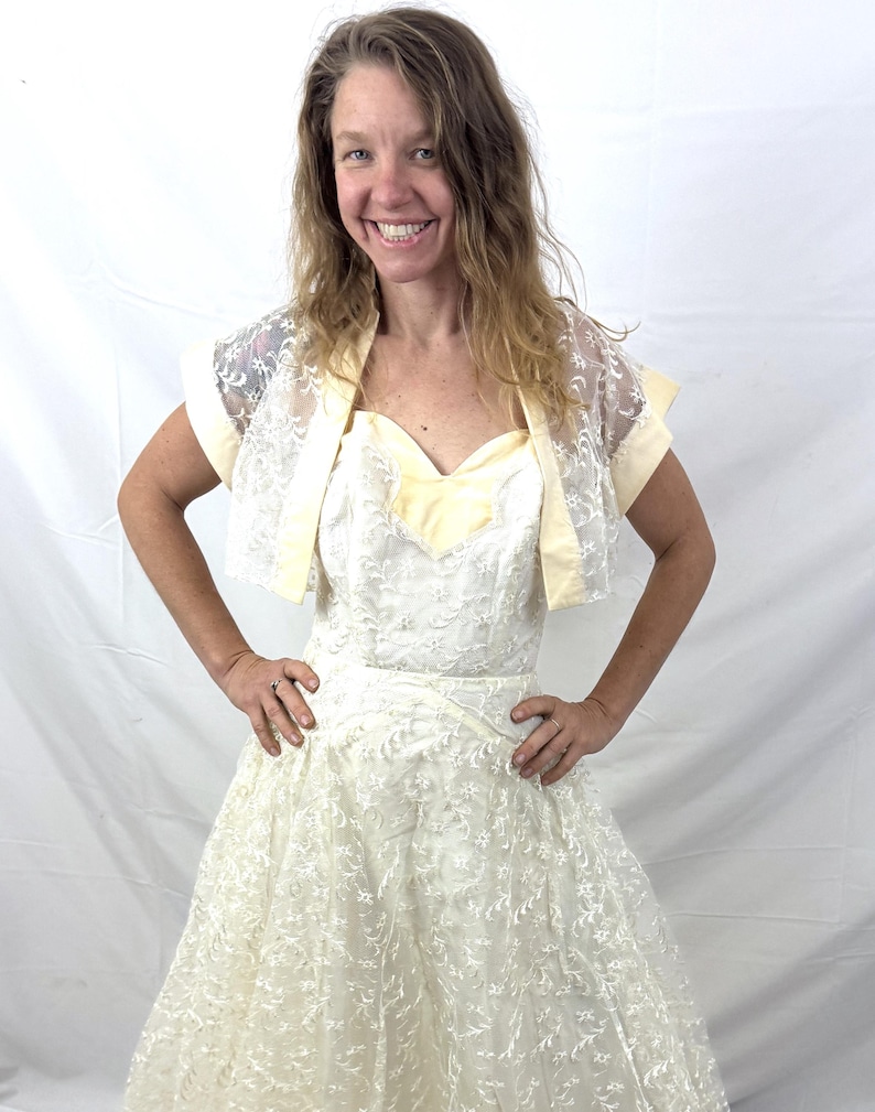 Puede incluir: Vestido de novia vintage de color marfil con una superposici&oacute;n de encaje y un escote coraz&oacute;n. El vestido presenta una falda amplia y se combina con un bolero de encaje a juego con ribetes amarillo p&aacute;lido. Un conjunto nupcial cl&aacute;sico.