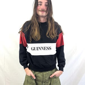 May include: Black sweatshirt with red shoulder accents and a white band across the chest. The word "GUINNESS" is printed in black on the white band. The person is wearing olive green cargo pants.