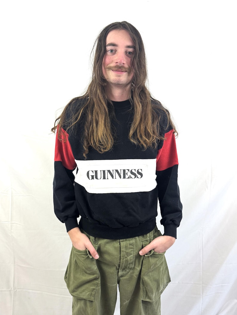 May include: Black sweatshirt with red shoulder accents and a white band across the chest. The word "GUINNESS" is printed in black on the white band. The person is wearing olive green cargo pants.