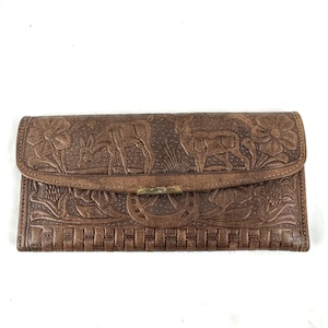May include: Brown leather wallet with embossed floral, deer, and horseshoe designs. The wallet features a woven pattern at the bottom and a metal clasp. The overall design is rustic and detailed.