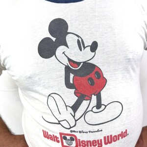 May include: White long-sleeve t-shirt featuring a smiling Mickey Mouse graphic in black, red, and white. The shirt has a navy blue neckline and the words "Walt Disney World" in red.