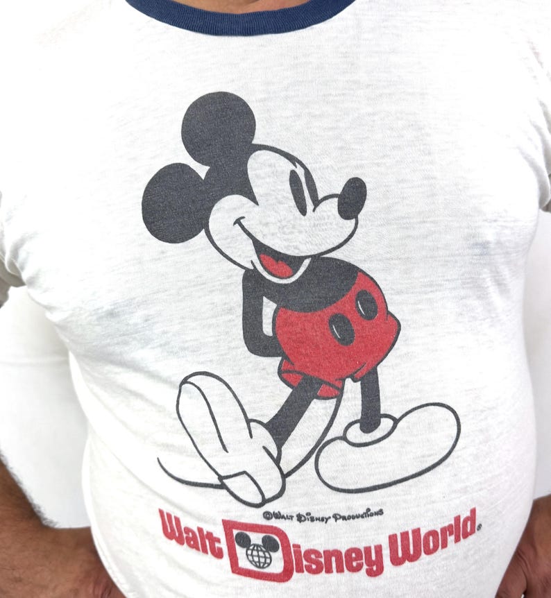 May include: White long-sleeve t-shirt featuring a smiling Mickey Mouse graphic in black, red, and white. The shirt has a navy blue neckline and the words "Walt Disney World" in red.