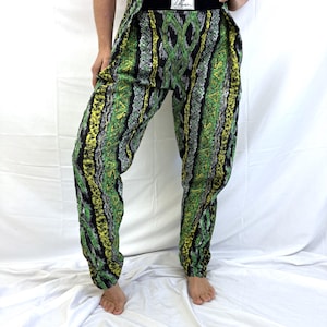 May include: Green, yellow, and black patterned pants with a wide black waistband. The pants have a unique design with a mix of geometric and organic shapes. The fabric appears to be lightweight, and the pants are gathered at the ankles.