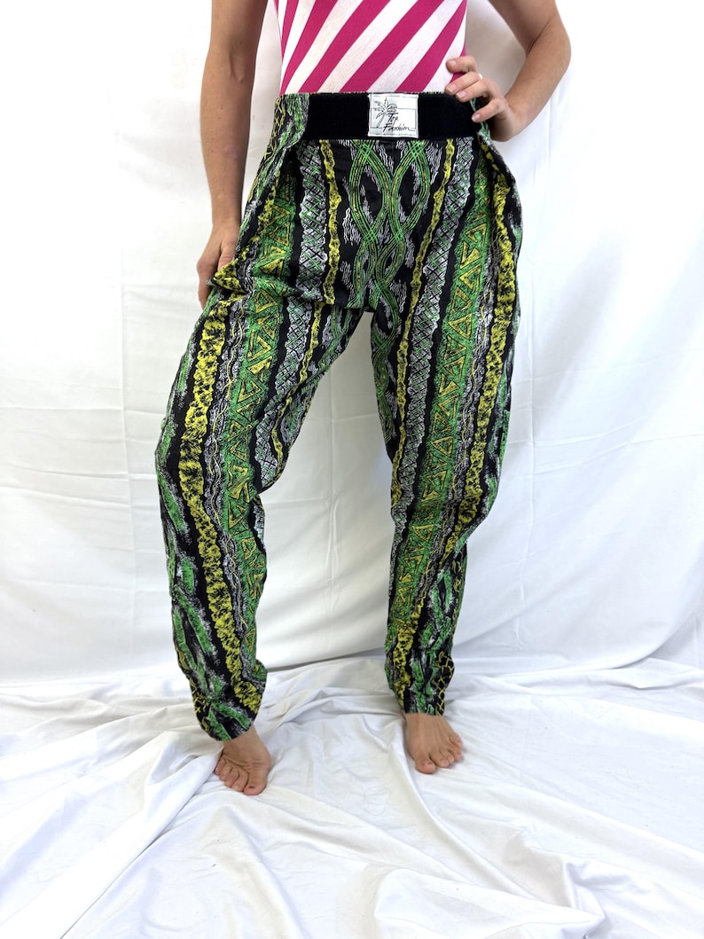 May include: Green, yellow, and black patterned pants with a wide black waistband. The pants have a unique design with a mix of geometric and organic shapes. The fabric appears to be lightweight, and the pants are gathered at the ankles.