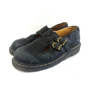 Puede incluir: Par de zapatos Mary Jane de cuero negro con dos correas de hebilla y una pala perforada. Los zapatos tienen una suela gruesa con costuras amarillas y hebillas doradas. El nombre de la marca es visible en las hebillas y en el lateral del zapato.