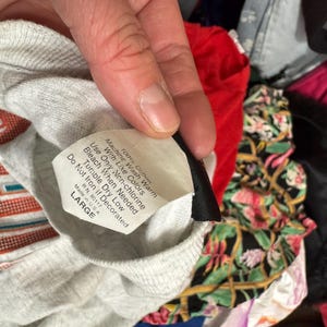 May include: A grey, long-sleeved shirt with a label stating "100% Cotton" and "LARGE". The shirt is surrounded by other garments, including a red top and a floral patterned fabric.