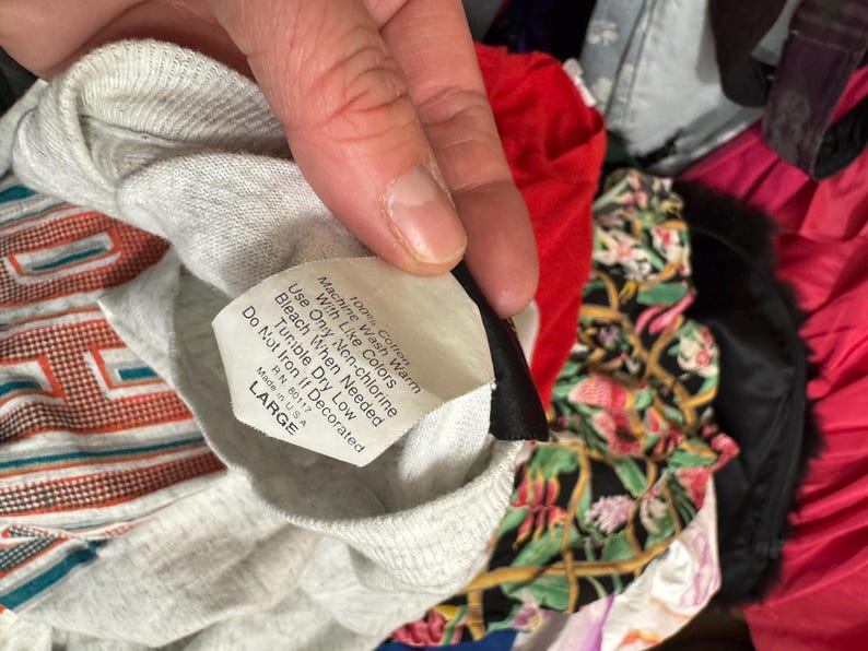 May include: A grey, long-sleeved shirt with a label stating "100% Cotton" and "LARGE". The shirt is surrounded by other garments, including a red top and a floral patterned fabric.
